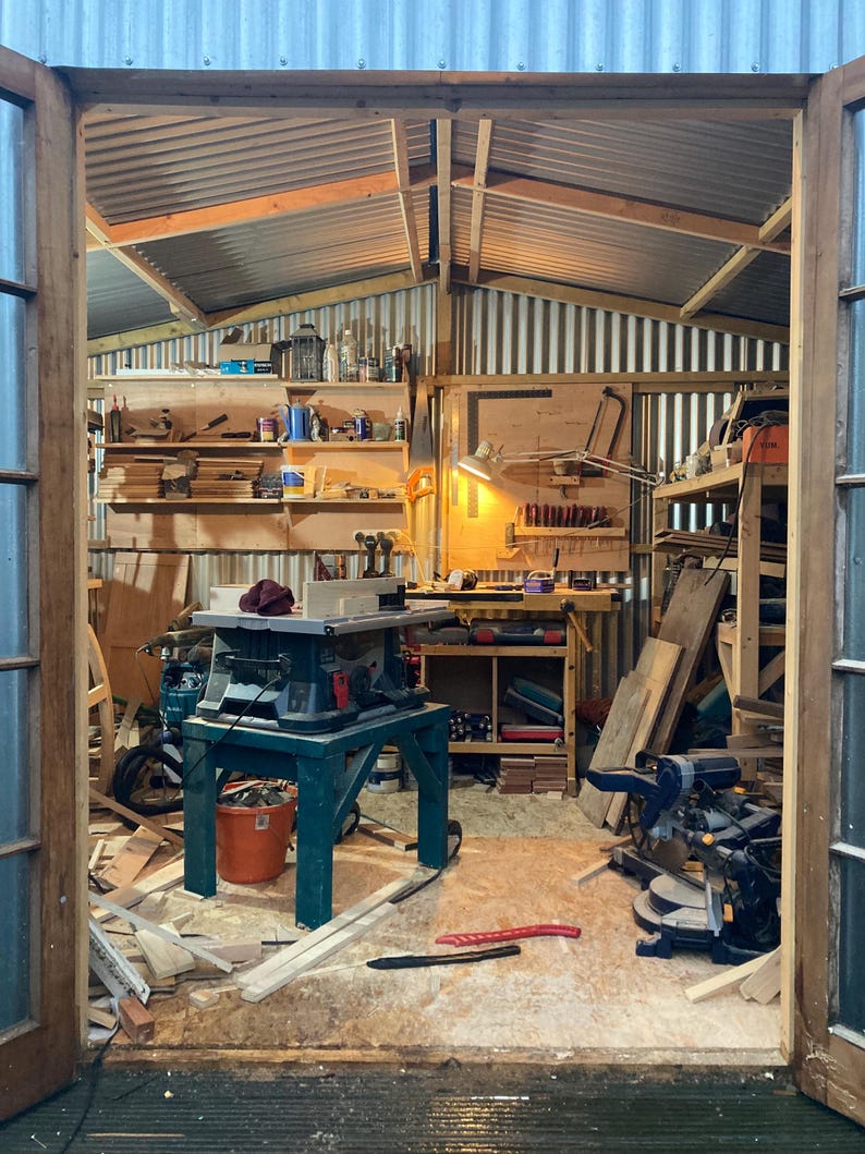 Puede incluir: Un taller desordenado con una sierra de mesa, una sierra de inglete y varias herramientas y materiales. El taller tiene un techo de metal y paredes de madera.