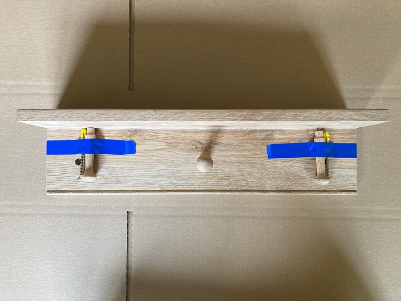 Puede incluir: Una estanter&iacute;a de madera con dos ganchos y un pomo en el centro. La estanter&iacute;a est&aacute; fijada a la pared con cinta de pintor azul.