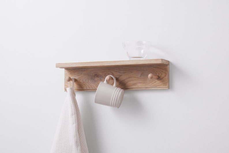 Puede incluir: Una estanter&iacute;a de madera con tres ganchos y un peque&ntilde;o cuenco de vidrio en la parte superior. Una toalla blanca cuelga de un gancho y una taza blanca cuelga de otro gancho.