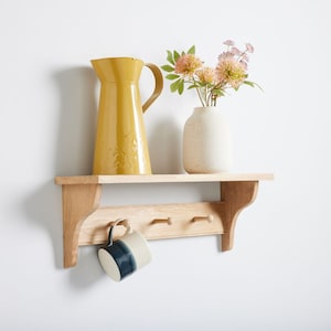 May include: A wooden shelf with three hooks, a yellow jug, a white vase with pink flowers, and a blue mug hanging from the hooks.