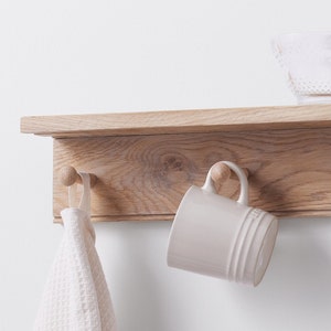 Puede incluir: Una estanter&iacute;a de madera con tres ganchos y una taza blanca colgada de uno de los ganchos. Una toalla blanca cuelga de otro gancho. La estanter&iacute;a est&aacute; montada en una pared blanca.