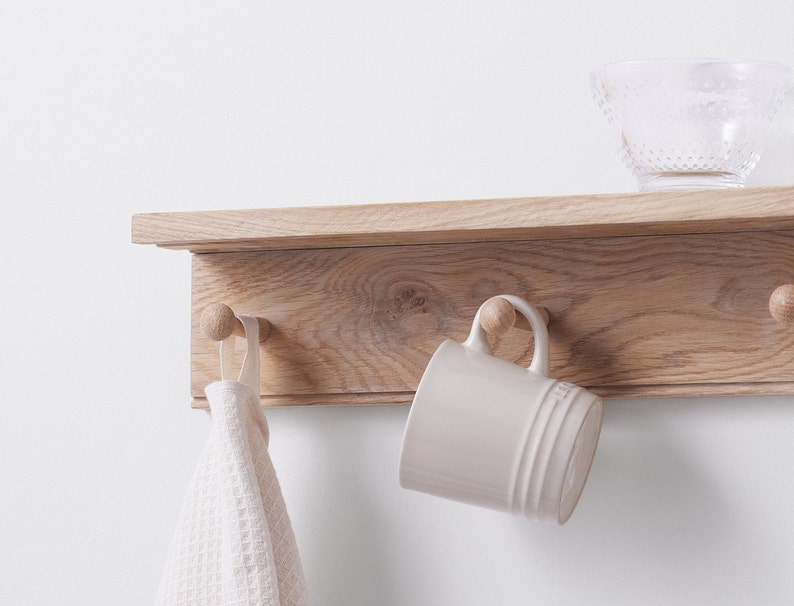 Puede incluir: Una estanter&iacute;a de madera con tres ganchos y una taza blanca colgada de uno de los ganchos. Una toalla blanca cuelga de otro gancho. La estanter&iacute;a est&aacute; montada en una pared blanca.