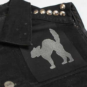 Könnte beinhalten: Eine schwarze Jeansweste mit silbernen Nieten am Kragen und einem schwarzen Katzen-Silhouetten-Patch auf der Brusttasche.