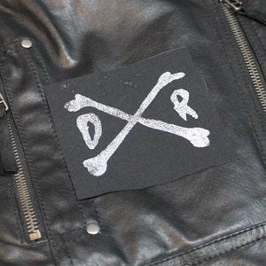 Könnte beinhalten: Schwarze Lederjacke mit Reißverschluss und einem schwarzen Patch mit weißen gekreuzten Knochen und den Buchstaben "D" und "R" auf der linken Brustseite.