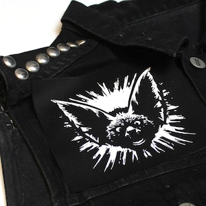 Könnte beinhalten: Schwarze Jeansweste mit silbernen Nieten und einem weißen Patch mit einer stilisierten Fledermaus mit einem Sternenausbruch-Design.