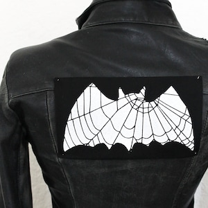 Könnte beinhalten: Schwarze Lederjacke mit einem schwarzen und weißen Fledermaus-Patch mit Spinnennetz-Design auf der Rückseite.