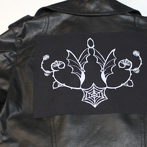 Könnte beinhalten: Schwarze Lederjacke mit einem schwarzen Stoff-Patch auf der Rückseite. Der Patch zeigt ein weiß gesticktes Design eines stilisierten Spinnennetzes mit Fledermausflügeln und floralen Akzenten.