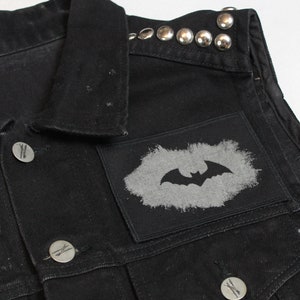Könnte beinhalten: Schwarze Jeansweste mit silbernen Nieten am Kragen und einem schwarzen und weißen Fledermaus-Patch auf der Vorderseite.