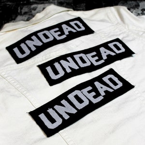 Könnte beinhalten: Drei schwarze Stoff-Patches mit dem Wort "UNDEAD" in weißen Buchstaben, auf eine weiße Jeansjacke genäht.