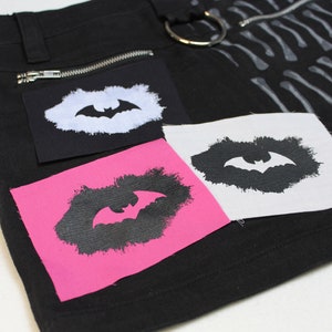 Könnte beinhalten: Ein schwarzer Jeansrock mit drei Patches. Die Patches sind weiß mit einer schwarzen Fledermaus-Silhouette, schwarz mit einer weißen Fledermaus-Silhouette und pink mit einer schwarzen Fledermaus-Silhouette.