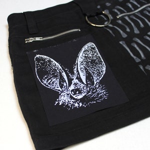 Könnte beinhalten: Schwarzer Minirock mit Reißverschlussdetail und einem weißen Fledermaus-Grafik-Patch. Der Rock hat ein Knochenmuster an der Seite.