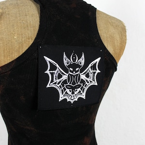 Könnte beinhalten: Schwarzes Tanktop mit einem schwarzen Patch, der eine weiße Fledermaus mit einem Spinnennetz-Design zeigt.