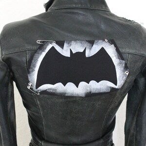 Könnte beinhalten: Schwarze Lederjacke mit einer weißen, bemalten Fledermaus-Silhouette auf dem Rücken. Die Fledermaus ist mit Sicherheitsnadeln befestigt.