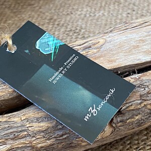 K&ouml;nnte beinhalten: Ein schwarzes Etikett mit wei&szlig;em Text, auf dem "Handmade - Primitive Jewelry Studio" und "mJ sencenik" steht. Das Etikett ist mit einer Schnur gebunden und liegt auf einem St&uuml;ck Treibholz.