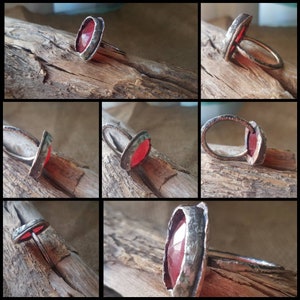 Pode incluir: Um anel de prata com uma pedra preciosa vermelha engastada em um engaste de bisel. O anel é mostrado de vários ângulos em uma superfície de madeira.
