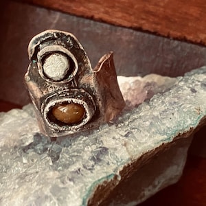 May include: A silver ring with two stones set in it. The stones are a round, white stone and a smaller, oval, brown stone. The ring is made of textured metal and has a unique, organic shape.