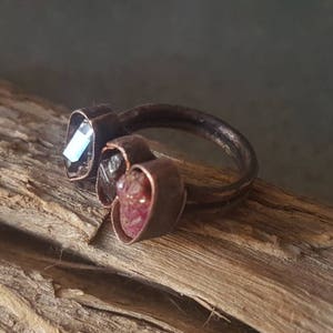Puede incluir: Un anillo de alambre de cobre con tres engastes en forma de corazón. Los engastes están llenos de pequeñas piedras de colores. El anillo está sobre una superficie de madera.