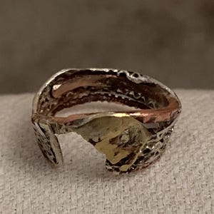May include: A handcrafted ring with a unique open design. The ring features a mix of metals, including silver, copper, and gold. The silver band has an intricate, textured pattern. The ring's open design adds a modern touch to the piece.