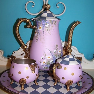 May include: A pink and gold tea set with a teapot, creamer, and sugar bowl. The set is decorated with a floral pattern and gold polka dots. The set is on a black and white checkered tray.