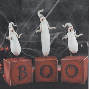 Pode incluir: Três figurinhas de fantasmas brancos com rostos pintados ficam em cima de três blocos de madeira vermelhos que soletram "BOO". Os blocos têm um desenho de teia de aranha preta e letras pretas.