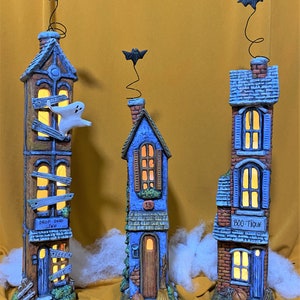 May include: Three ceramic Halloween houses with lit windows. The houses are painted in blue, brown, and red with black bats on top. The houses have signs that read "Drop Dead Inn", "Boo-Tique", and "13".