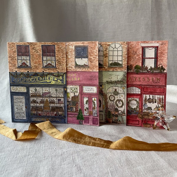 May include: A set of four illustrated Christmas cards featuring storefront scenes. The cards are decorated with festive greenery, wreaths, and a variety of holiday details. The storefronts are labelled "Chocolatier", "Art Gallery", "Local Crafts", and "Atessein".