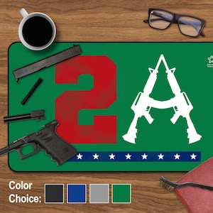 May include: Green gun cleaning mat with a red number "2" and two white guns forming the letter "A". The mat has a row of white stars along the bottom. The text "Liberty Defender" is in the top right corner.