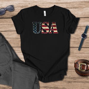 May include: Black t-shirt featuring the word "USA" in a distressed design, incorporating the stars and stripes of the American flag. The image includes a pair of grey jeans, sunglasses, a watch, and a brown belt. The shirt is a casual wear item.