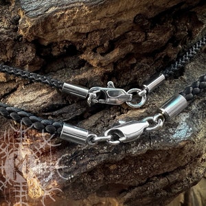 May include: Two black braided leather cords with silver clasps. The cords are lying on a piece of rough, brown wood.
