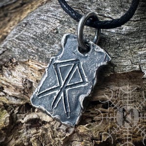 Könnte beinhalten: Eine silberfarbene Halskette mit Anhänger und schwarzem Band. Der Anhänger hat eine raue, strukturierte Oberfläche und zeigt ein geschnitztes geometrisches Symbol. Die Halskette wird vor einem strukturierten Holzgrund gezeigt. Der Text "MAGIKREBEL" ist unten sichtbar.