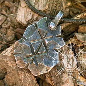 Pode incluir: Pingente de metal texturizado feito à mão com um design geométrico e um cordão de couro. O pingente tem uma aparência escura e desgastada, com linhas gravadas formando um padrão de estrela. A palavra "MAGICREBEL" é visível na parte inferior.