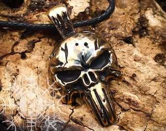 Pode incluir: Um pingente de metal dourado em forma de caveira com um design detalhado. O pingente está pendurado em um cordão preto.