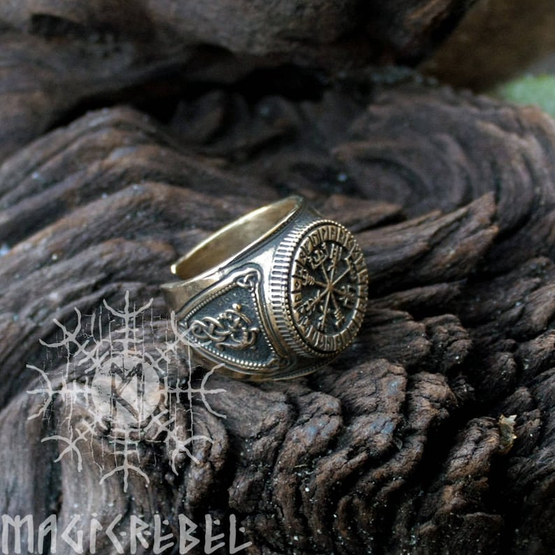 May include: A silver and gold ring with a compass design and runes around the edge. The ring is on a dark brown wooden surface.