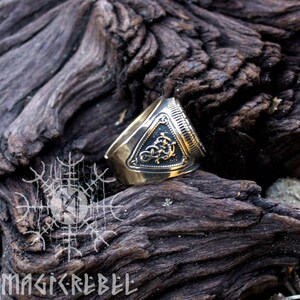 May include: A gold-toned ring with a triangular design featuring intricate details and a raised inscription. The ring is set against a rustic wooden background.