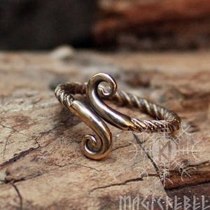 May include: A bronze ring with a twisted band and a stylized design of two curved shapes. The ring is on a wooden surface.