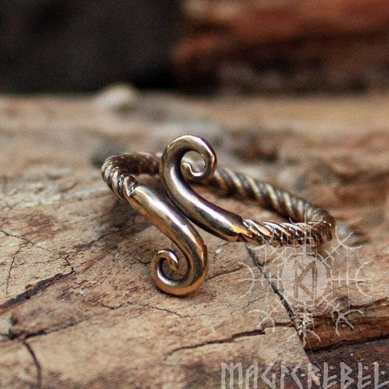 May include: A bronze ring with a twisted band and a stylized design of two curved shapes. The ring is on a wooden surface.