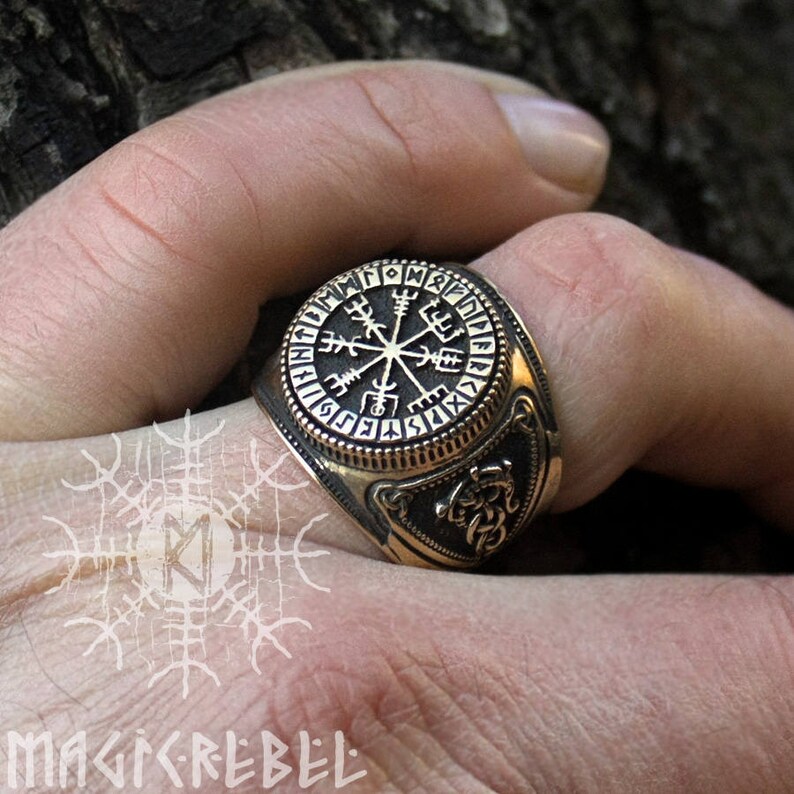 May include: A gold-toned ring with a circular design featuring a compass rose and runes. The ring has a raised edge and a textured band.