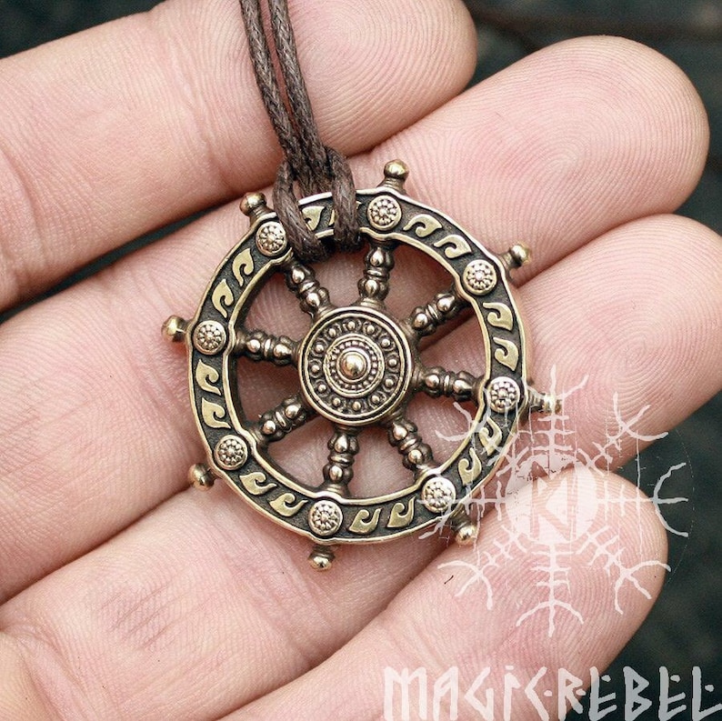 Puede incluir: Colgante de collar de color bronce con un dise&ntilde;o de rueda detallado con patrones intrincados y un elemento circular central. El colgante est&aacute; suspendido de un cord&oacute;n marr&oacute;n. Es una joya.
