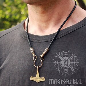 May include: A black leather cord necklace with a gold-toned pendant featuring a detailed hammer design. The pendant has a decorative pattern of small circles and a loop at the top. The necklace has two decorative clasps.