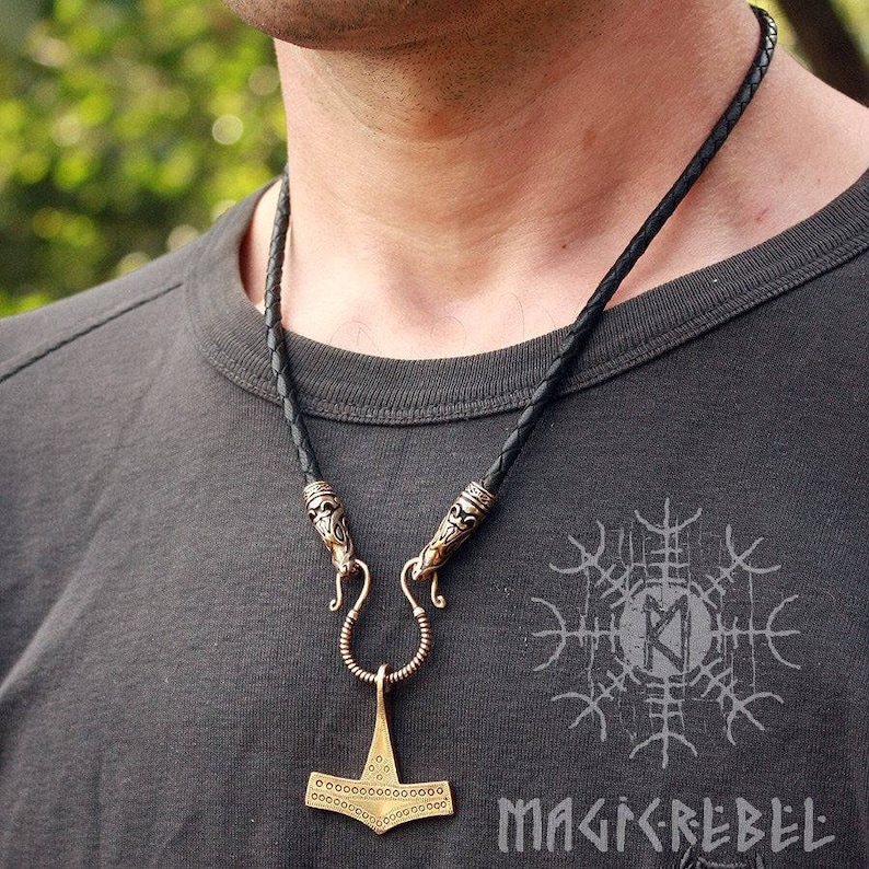 May include: A black leather cord necklace with a gold-toned pendant featuring a detailed hammer design. The pendant has a decorative pattern of small circles and a loop at the top. The necklace has two decorative clasps.