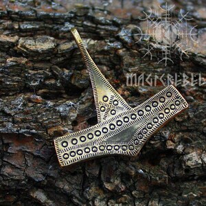 May include: A gold-colored metal pendant with a geometric design. The pendant is shaped like a stylized hammer with a circular pattern around the edges.