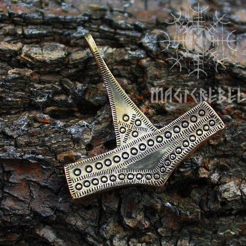 May include: A gold-colored metal pendant with a geometric design. The pendant is shaped like a stylized hammer with a circular pattern around the edges.