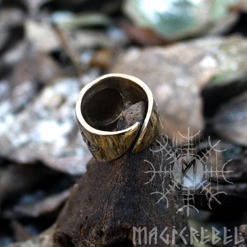 May include: A bronze ring with a textured surface and a raised design. The ring is sitting on a piece of wood with a pattern of runes.