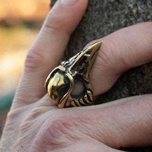 Op de afbeelding: Een ring van goudkleurig metaal in de vorm van een vogelkop met een gedetailleerd ontwerp. De ring wordt aan een vinger gedragen.