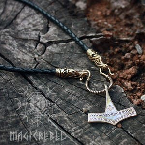 May include: A gold-toned Mjolnir pendant necklace with a braided black leather cord. The pendant features a detailed design with a circle pattern around the edges. The necklace has a clasp with a dragon head design.