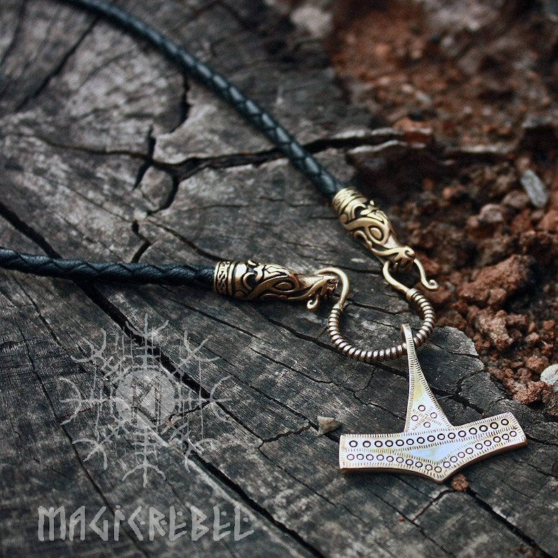 May include: A gold-toned Mjolnir pendant necklace with a braided black leather cord. The pendant features a detailed design with a circle pattern around the edges. The necklace has a clasp with a dragon head design.