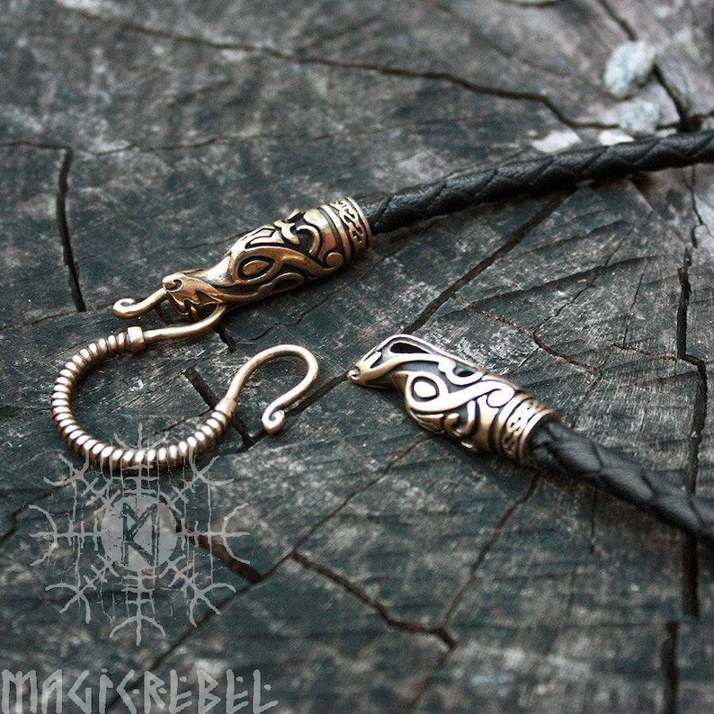 May include: A black leather cord necklace with two ornate gold-toned metal dragon head clasps. The clasps have intricate details and a Celtic knot design.