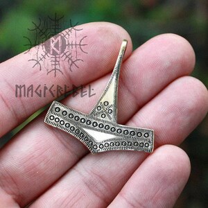 May include: A silver-colored metal pendant in the shape of a hammer with a decorative pattern of small circles around the edges. The pendant is held in a hand.