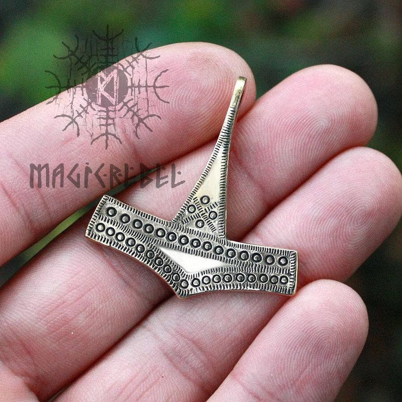 May include: A silver-colored metal pendant in the shape of a hammer with a decorative pattern of small circles around the edges. The pendant is held in a hand.