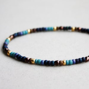 Pode incluir: Uma pulseira fina de contas com um padrão repetitivo de contas mate em azul escuro, azul claro e turquesa, intercaladas com pequenos espaçadores metálicos dourados e contas castanho-escuras facetadas num design circular contínuo.